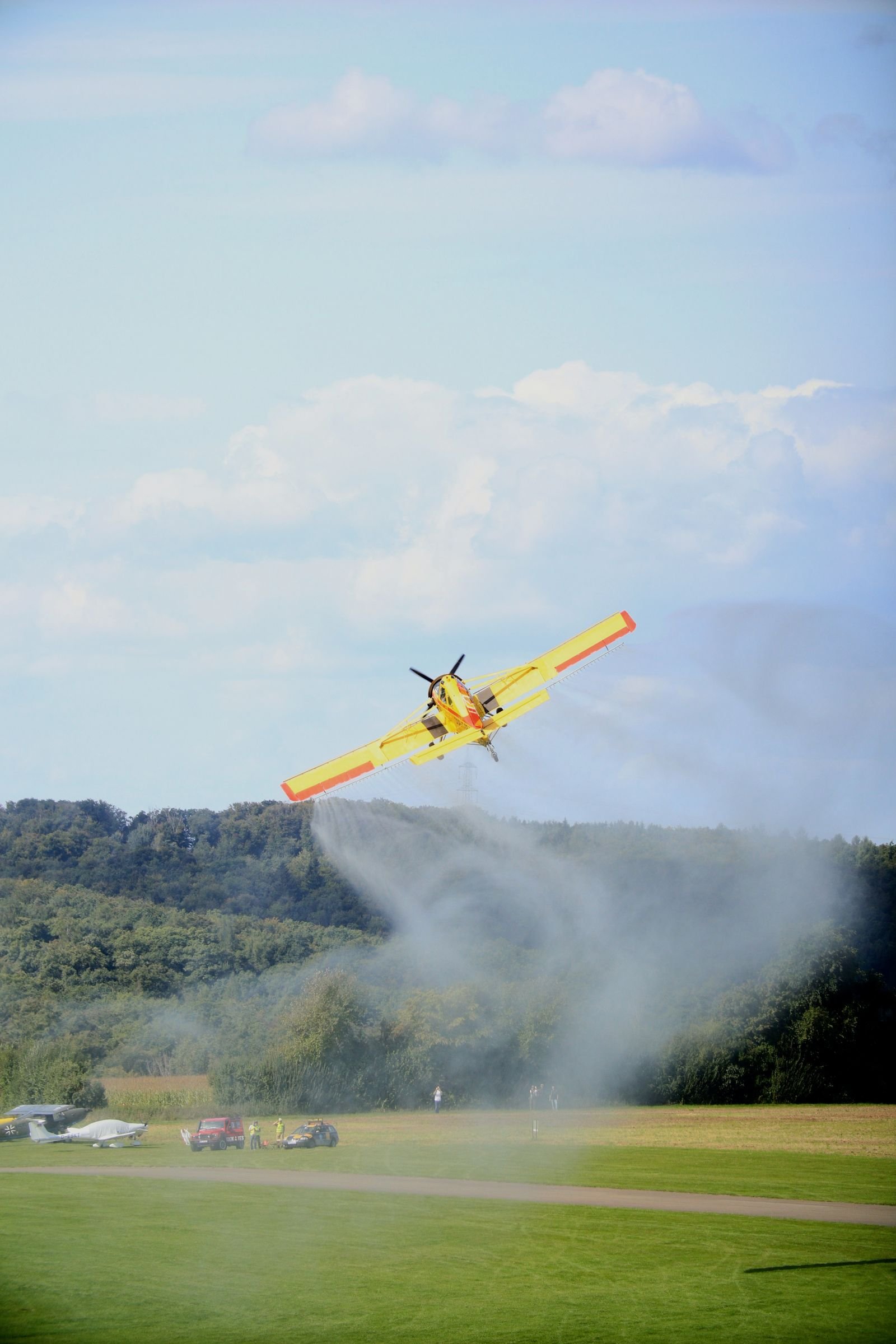Pesticide drift exposure: yellow crop duster plane spraying a green field, leaving a trail of mist