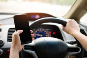 Distracted driving deaths: hand holding a smartphone while gripping a steering wheel, showing distracted driving
