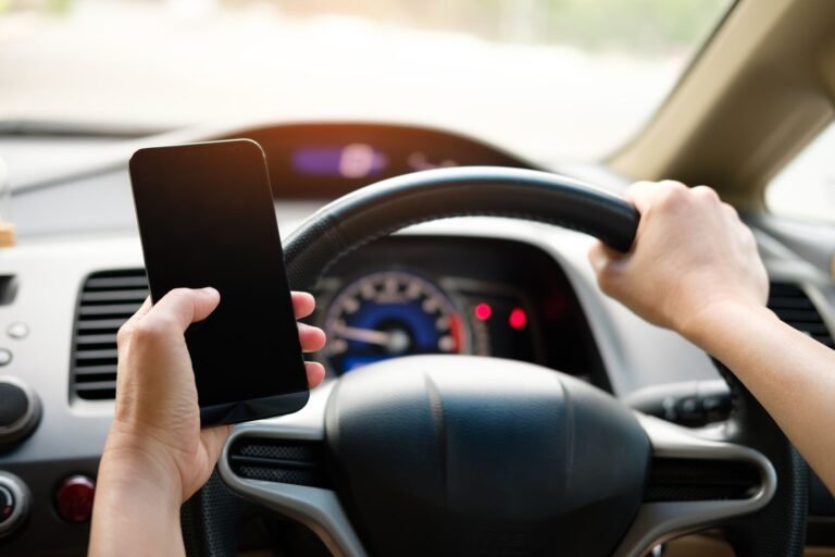 Distracted driving deaths: hand holding a smartphone while gripping a steering wheel, showing distracted driving