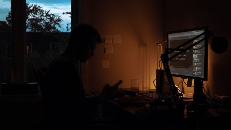 Warning signs a child is targeted online: silhouette of an adult using a phone beside a glowing computer screen in a dark room