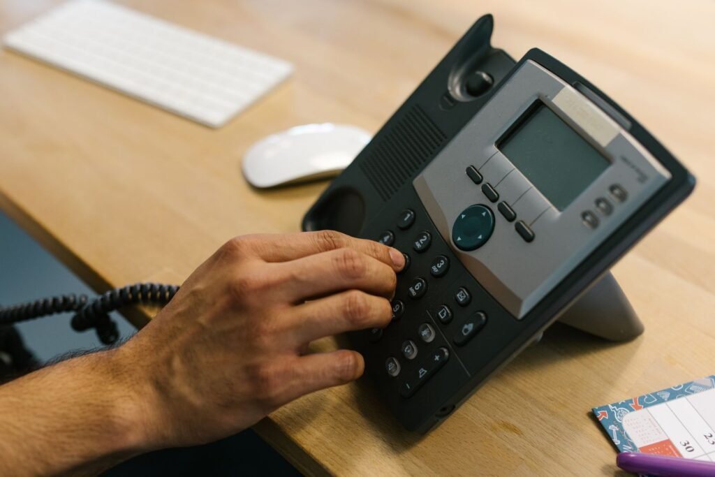 Hand dialing a desk phone beside a keyboard and planner in an office