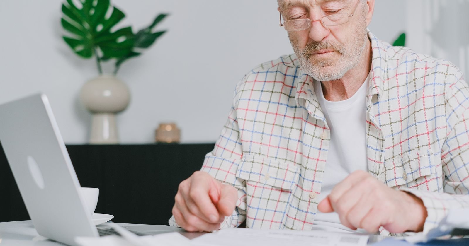 Older man at home reviewing final expense claim documents and paperwork at a table with a laptop