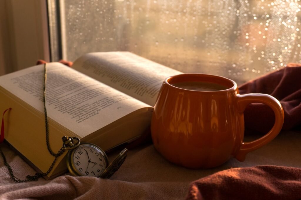 Mug, open book and pocket watch by a window on a rainy autumn day, a quiet moment of self-care