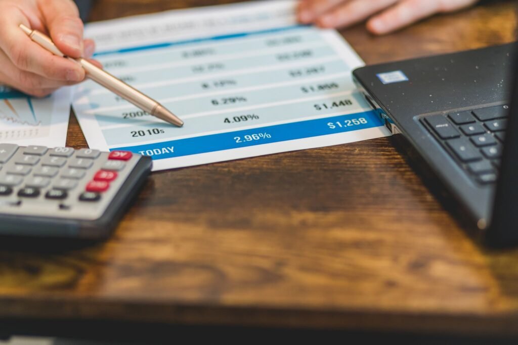 Hand with a pen reviewing figures on paper next to a calculator and laptop